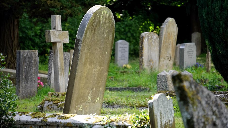 The image displays a close-up view of several weathered headstones in a cemetery, with the foreground featuring a large, upright stone with visible moss and lichen growth along its edges. Surrounding 